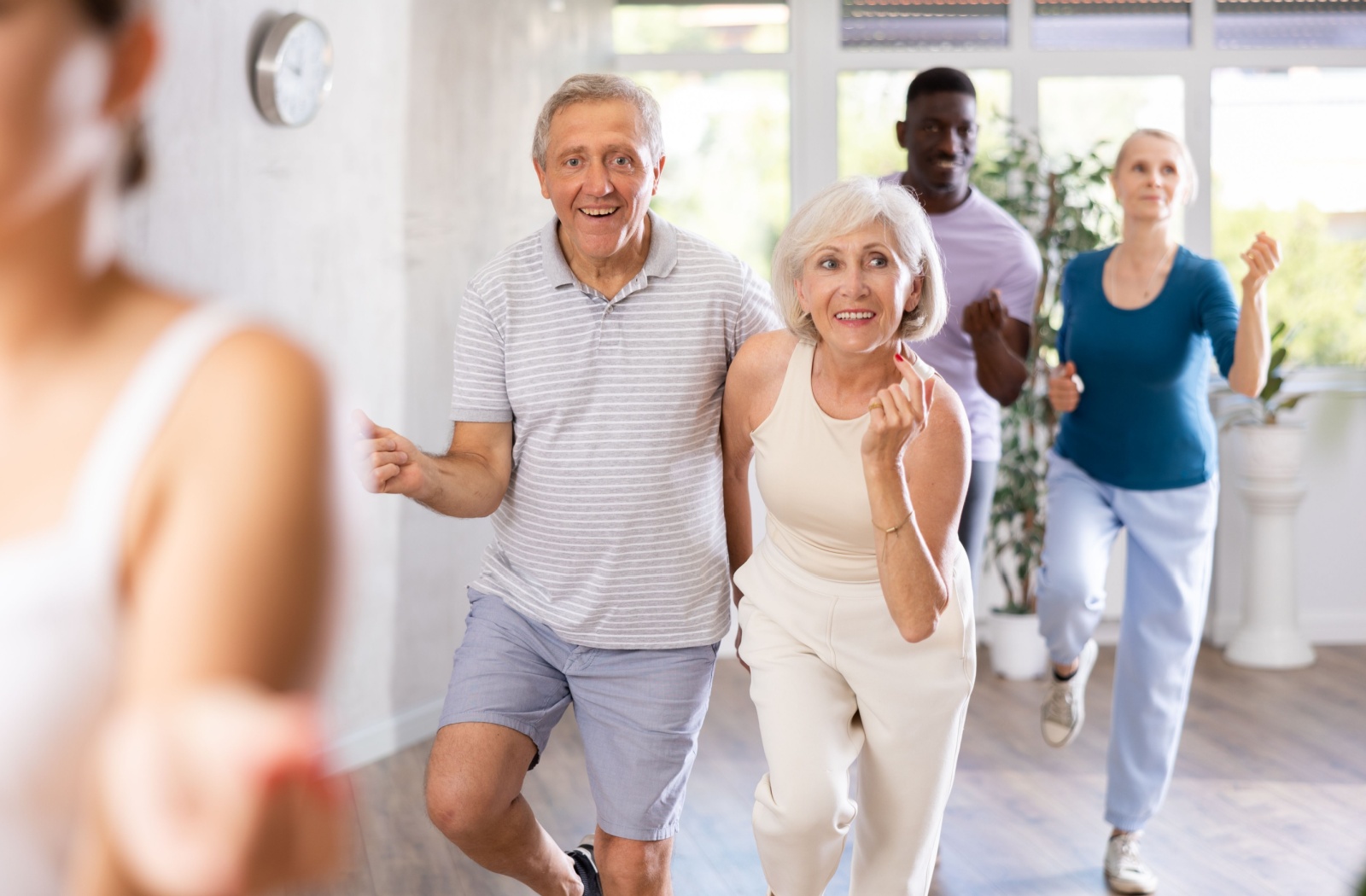 A group of smiling seniors is participating in a light dance class in a bright room, moving forward with raised hands.