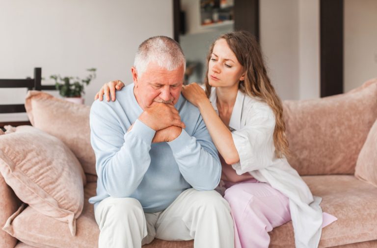 An adult child tries to calm their stressed senior parent as they both sit on a couch.