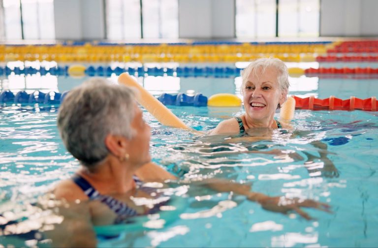 Two smiling seniors are in a swimming pool, one looking at the other and supporting their body with an orange foam pool noodle, during a water exercise session.